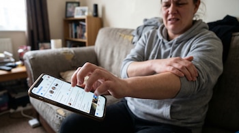 Mujer sentada en un sofá con expresión de dolor, sosteniendo un celular y sujetándose el brazo, mostrando incomodidad por su uso.