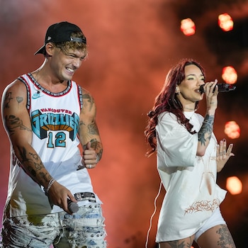 Un hombre y una mujer cantan en un escenario. Él usa gorra, camiseta de básquet y jeans rotos. Ella tiene el cabello rojo, camiseta y shorts blancos