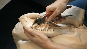 Primer plano de manos doblando cuero alrededor de una horma de zapato, asegurándolo con clavos pequeños usando pinzas