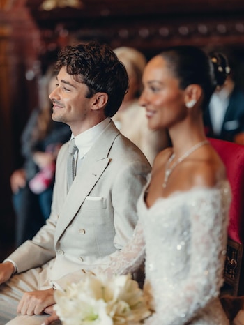 Charles Leclerc sonriendo, vistiendo un traje beige, junto a Alexandra Saint Mleux, difuminada, con un vestido de novia blanco y un ramo floral