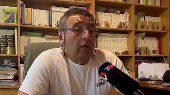 Un hombre con anteojos y camiseta blanca habla frente a un micrófono ante una biblioteca con libros y carpetas
