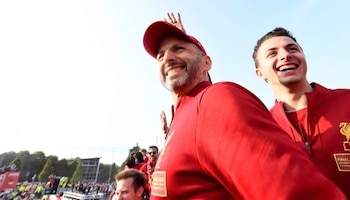 Un hombre con gorra roja y chaqueta deportiva sonríe y saluda con la mano levantada junto a otro joven con chaqueta del Liverpool FC