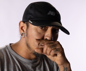 Retrato de un hombre con gorra negra, camiseta gris, barba, expansor en la oreja izquierda y tatuajes visibles en los dedos de la mano derecha