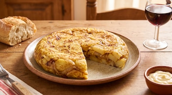 Tortilla de papas con cebolla cortada en un plato, pan, copa de vino tinto, mayonesa en cuenco y cubiertos sobre mesa de madera.