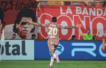 Jugador de fútbol con camiseta número 20, Alex Valera, corre con los brazos abiertos en el césped de un estadio frente a una tribuna con banderas