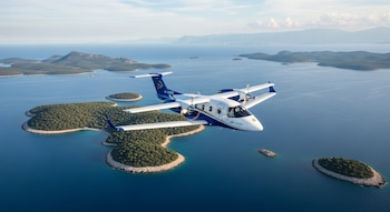 Un avión blanco, azul y dorado con dos hélices vuela sobre el mar azul con varias islas verdes con playas rocosas en un día soleado.
