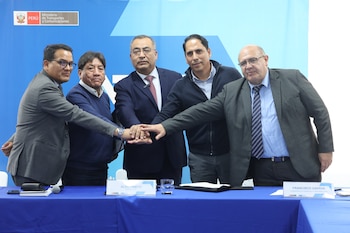 Cinco hombres con vestimenta formal y casual apilan sus manos sobre una mesa azul durante la firma de un acuerdo, con el logo del Ministerio de Transportes y Comunicaciones al fondo