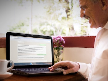 Hombre sonriente de edad avanzada con camisa blanca usa una netbook negra sobre una mesa de madera, junto a una taza blanca y flores púrpuras en un jarrón