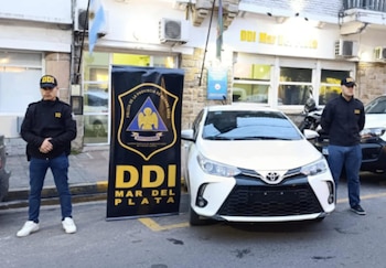 Dos hombres con uniformes de la DDI y gorras posan junto a un Toyota Yaris blanco y un cartel con el logo de DDI Mar del Plata, frente a un edificio
