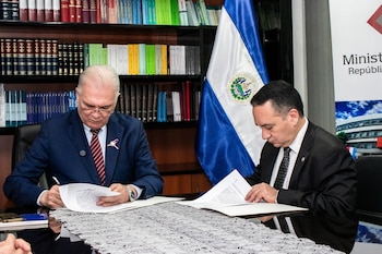 Dos hombres en traje, uno de ellos con corbata a rayas y una escarapela, firman documentos en un escritorio frente a una bandera de El Salvador y estanterías de libros