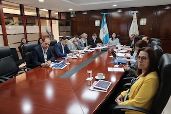 Once personas sentadas en una sala de conferencias con una mesa larga, documentos, laptops, vasos y tazas. Se ven dos banderas, una de Guatemala