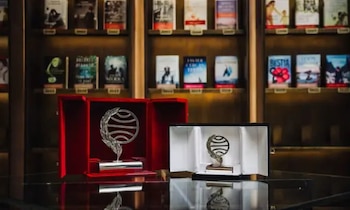 Dos esculturas metálicas con forma de globo y rama en estuches abiertos sobre una mesa de vidrio con libros de fondo