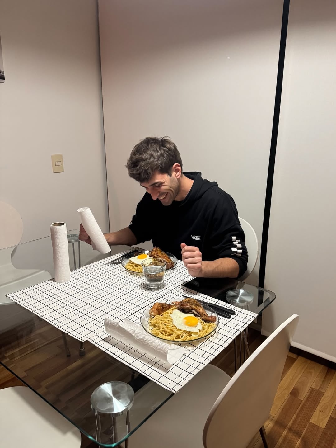 La pareja del youtuber sonríe mientras cena un plato de pasta con pollo y huevo frito en una mesa de cristal con un mantel a cuadros