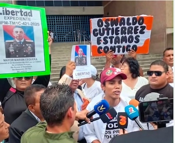 Varias personas sostienen pancartas con fotos y nombres ante micrófonos de prensa frente a la escalera de un edificio