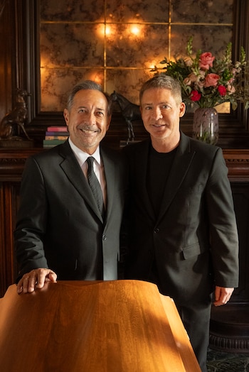 Dos hombres con trajes oscuros, uno con corbata, sonríen frente a una chimenea con espejo, luces, un ramo de flores y figuras decorativas