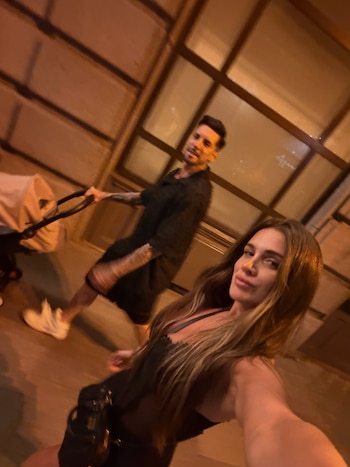 Una mujer con cabello largo oscuro en primer plano se toma una selfi, mientras un hombre con tatuajes empuja un cochecito de bebé detrás de ella por la noche