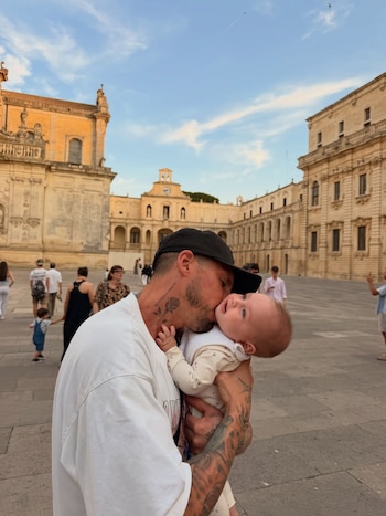 Hombre con gorra negra y tatuajes besa la mejilla de un bebé. Están en una plaza exterior con edificios clásicos de color beige y personas al fondo bajo el cielo