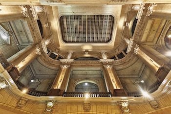 Vista contrapicada del interior de un edificio con columnas de piedra, balaustradas, arcos, lámparas encendidas y una claraboya rectangular