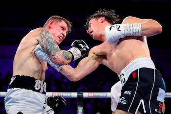 Dos boxeadores en el ring. El de la izquierda, con guantes blancos y tatuajes, recibe un puñetazo del boxeador de la derecha, quien usa guantes negros