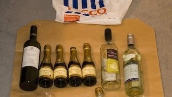 Una botella de vino tinto, tres botellas pequeñas de champán, y dos botellas de vino blanco sobre una superficie clara, con una bolsa Tesco al fondo