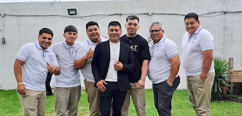 Seis hombres sonríen mientras posan al aire libre frente a una pared blanca y pasto verde. Cinco visten polos blancos con el logo 'H.T.', uno lleva camiseta negra 'H.T.'