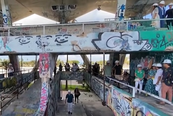 Edificio de concreto con graffiti en columnas y paredes, personas con cascos blancos en pasillos y escaleras, vegetación visible al fondo