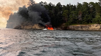 Mar, costa rocosa con árboles, embarcación blanca incendiándose con llamas y humo negro, cielo con nubes anaranjadas a la izquierda.