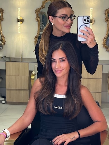 Antonela Roccuzzo, con cabello castaño largo y liso, sonríe a la cámara en un salón, mientras una estilista con gafas se toma una selfie detrás de ella