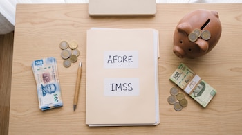 Una carpeta con etiquetas de AFORE e IMSS, billetes de pesos mexicanos, monedas, un bolígrafo y una alcancía sobre una mesa de madera.