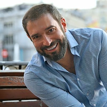 Hombre de mediana edad con barba oscura y sonrisa amplia, lleva camisa azul con estampado sutil, sentado en un banco de madera con fondo urbano difuminado