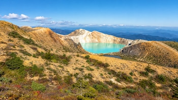 Lago de color turquesa dentro de un cráter rocoso, rodeado por laderas de tierra amarillenta con vegetación baja bajo un cielo azul