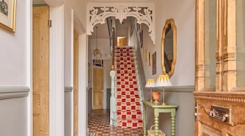 Un pasillo iluminado con piso de baldosas de colores, escaleras con alfombra roja, una mesa auxiliar, dos lámparas, un espejo y varias puertas de madera.