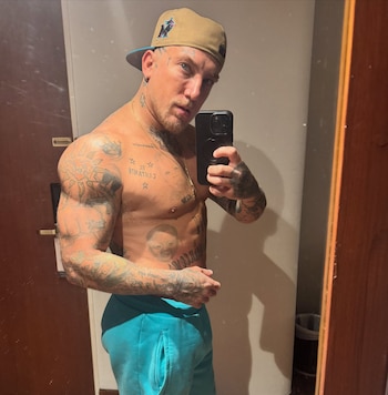 Un hombre sin camiseta, con gorra, tatuajes y pantalones cortos turquesa, se toma una selfie frente a un espejo con un teléfono móvil
