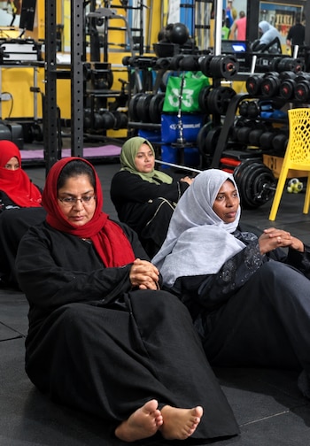 Cuatro mujeres con túnica y velo permanecen sentadas con las piernas flexionadas en el suelo de un gimnasio con pesas al fondo