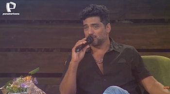 Primer plano de Pablo Heredia hablando en un micrófono con un fondo de madera. Viste una camisa oscura y un collar. Logotipo de Panamericana TV arriba a la izquierda
