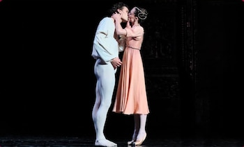 Un bailarín con vestimenta blanca y una bailarina con vestido rosado se besan sobre un escenario oscuro