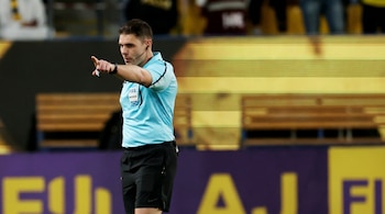 Un árbitro de fútbol masculino, con camiseta azul claro y pantalón corto negro, señalando con el brazo derecho en un estadio de fútbol.