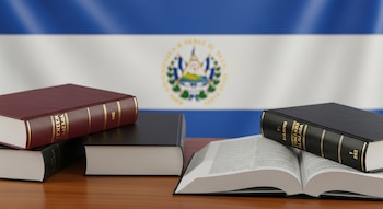 Tres libros, uno rojo y dos negros, sobre un escritorio de madera; uno de los libros negros está abierto mostrando texto, con la bandera de El Salvador difuminada al fondo.