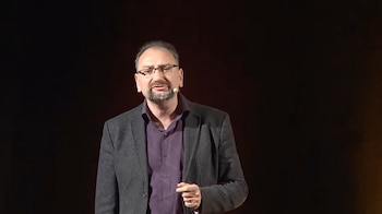 Un hombre de mediana edad con gafas, barba y camisa morada bajo un blazer oscuro, habla a un micrófono de solapa en un escenario con fondo oscuro
