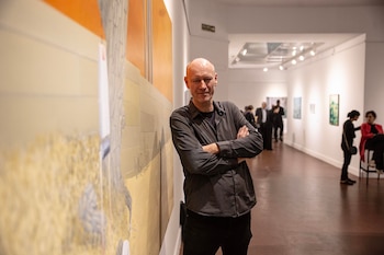 Hombre calvo con camisa oscura de manga larga y pantalones negros, brazos cruzados, apoyado en una pared junto a una gran obra de arte abstracta en una galería