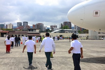 Grupo de personas, mayormente hombres, camina por una pista de aterrizaje junto a la parte frontal de un avión blanco con edificios urbanos al fondo