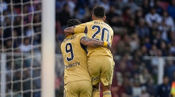 Vista trasera de dos futbolistas con camisetas doradas, Jiménez (9) y Velasco (20), abrazándose en el campo. Red de portería y multitud borrosa en el fondo.