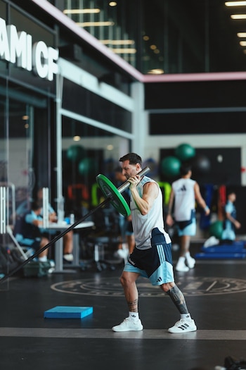 Lionel Messi, con camiseta blanca y shorts, levanta una barra con disco verde en un gimnasio. Se observan otros deportistas y equipamiento deportivo