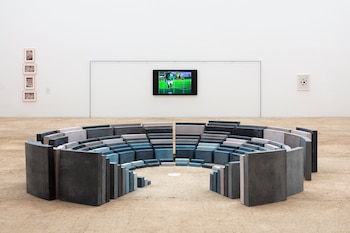 Instalación circular de bloques de hormigón grises y azules sobre suelo de arena clara, con pantalla de televisión mostrando partido de fútbol y cuadros en la pared