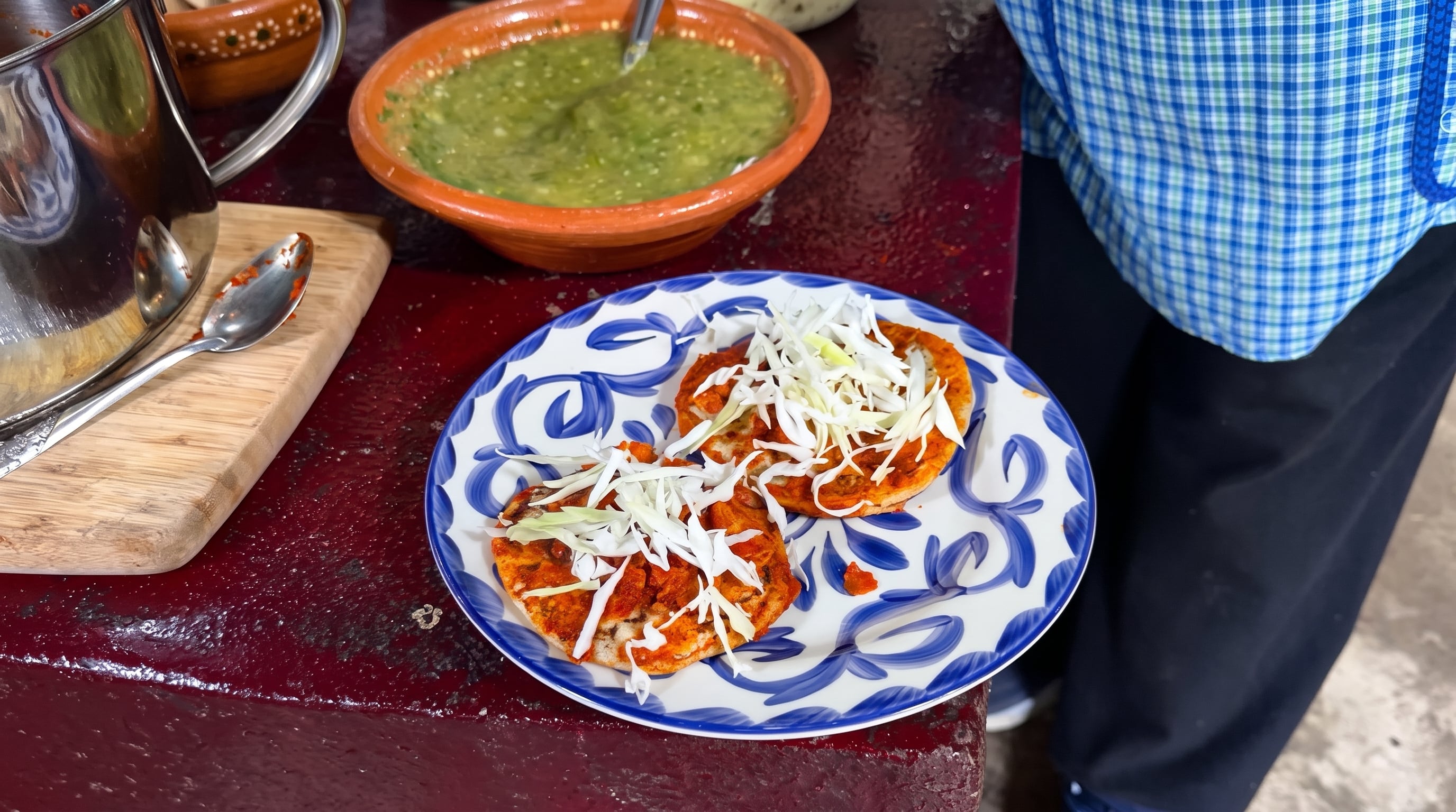 Un plato decorado contiene dos tortillas preparadas con salsa roja y repollo picado, al lado de un recipiente de barro con salsa verde. (Captura de pantalla De mi Rancho a tu Cocina)