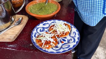 Dos tortillas redondas con salsa roja y repollo sobre un plato blanco y azul, junto a un recipiente de barro con salsa verde y una olla.