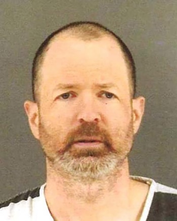 Un hombre de mediana edad con cabello corto y barba, mirando al frente en una foto policial, con un fondo liso