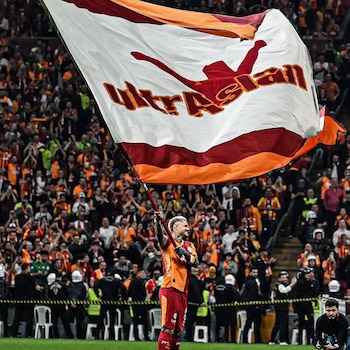 Mauro Icardi, con camiseta naranja y roja del Galatasaray, sostiene una gran bandera blanca, roja y naranja frente a la multitud en un estadio de fútbol