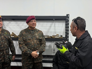 Dos militares en uniforme camuflado escuchan a un técnico con guantes de trabajo frente a estantes metálicos con pistolas en estuches