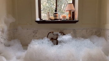 Bañera de mármol con abundante espuma. Grifo de bronce. Ventana con follaje exterior. En el alféizar, dos botellas, un cepillo, una vela encendida y una lámpara.
